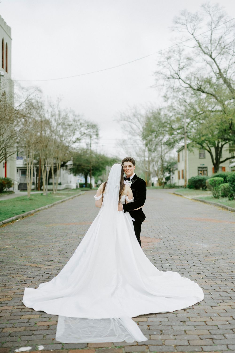 Elegant bride in flowing white gown twirls on cobblestone street during wedding photoshoot.
