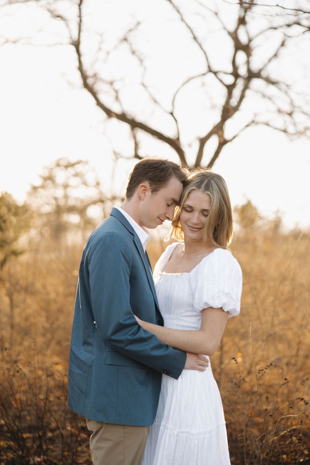 A romantic couple embraces in a golden field at sunset with bare trees in the background.