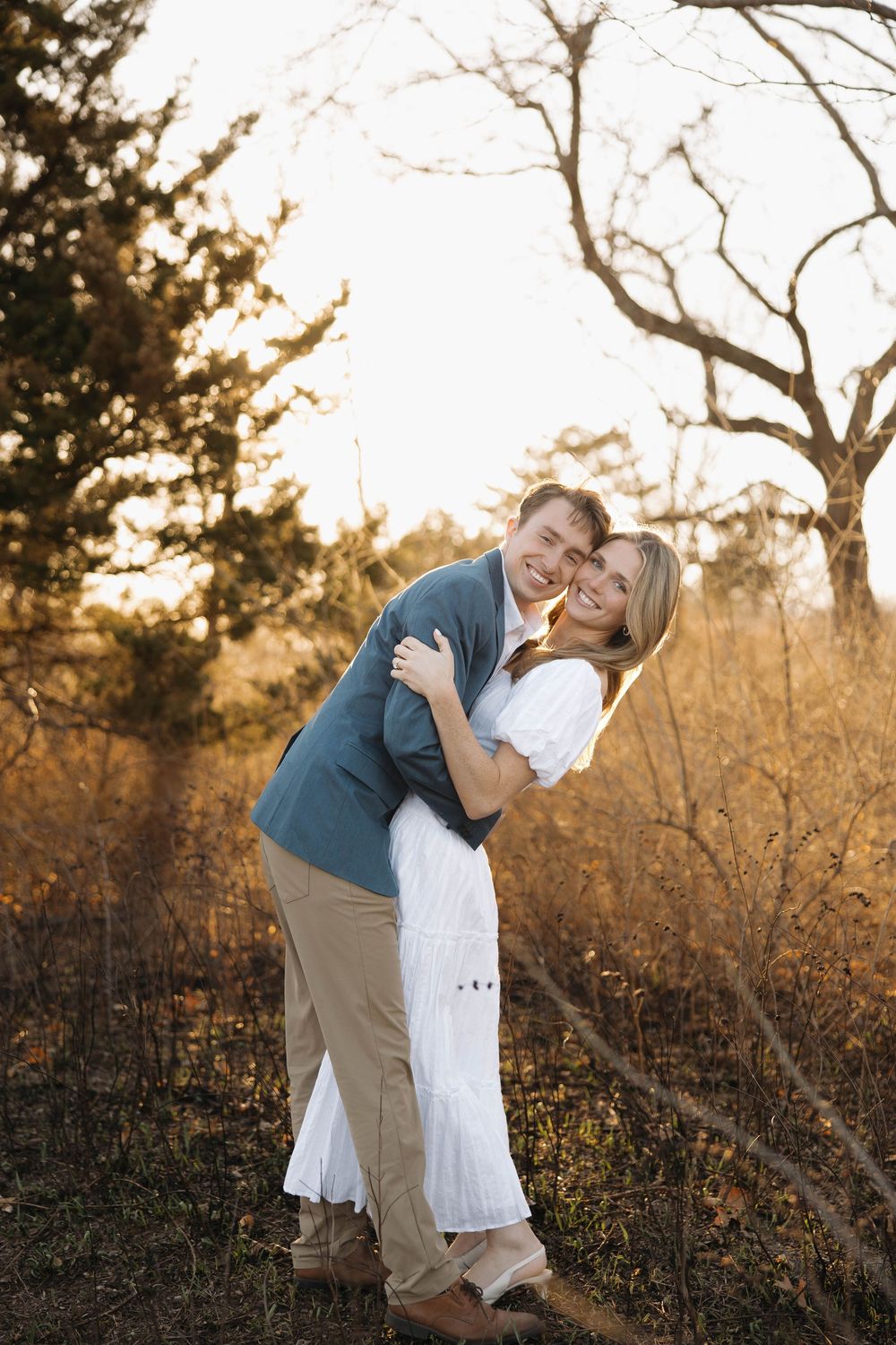 A couple embraces in a romantic outdoor photoshoot at sunset with golden light filtering through bare tree branches.