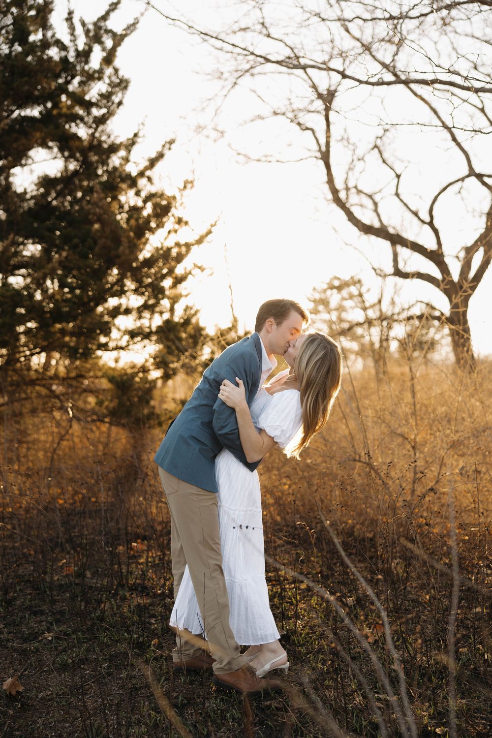 A couple embraces in a romantic outdoor setting during golden hour with bare winter trees in the background.