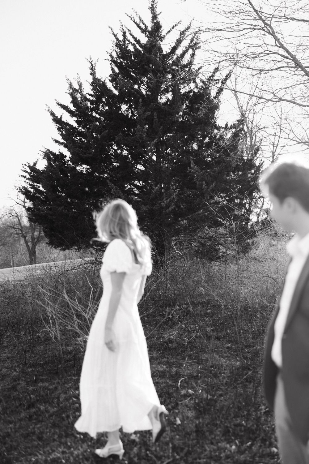 A black and white photograph shows two people outdoors near an evergreen tree on a hillside.