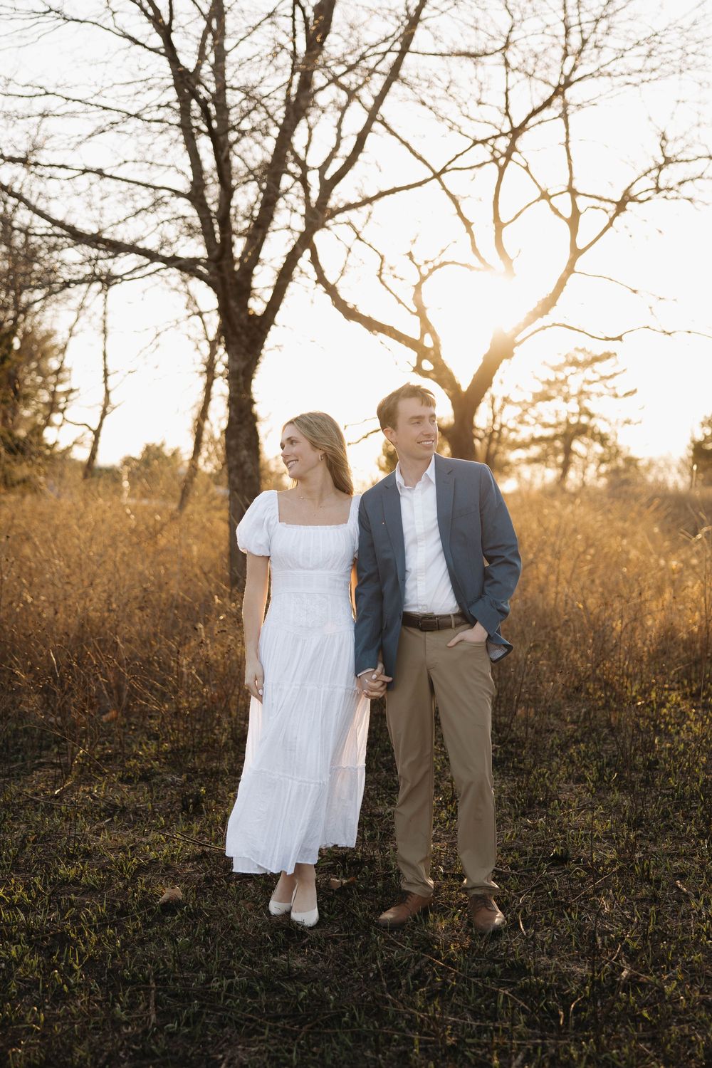 A couple in white dress and suit walk through a golden field at sunset as bare winter trees frame the scene.