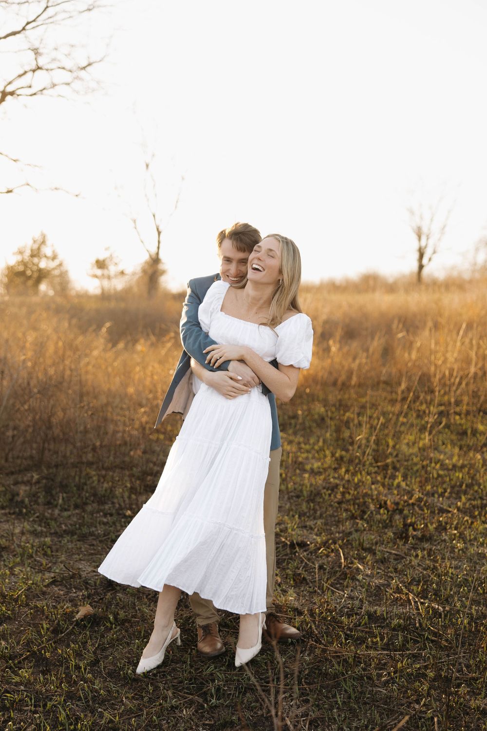 A couple in white clothing dances together in a golden sunlit field during sunset.