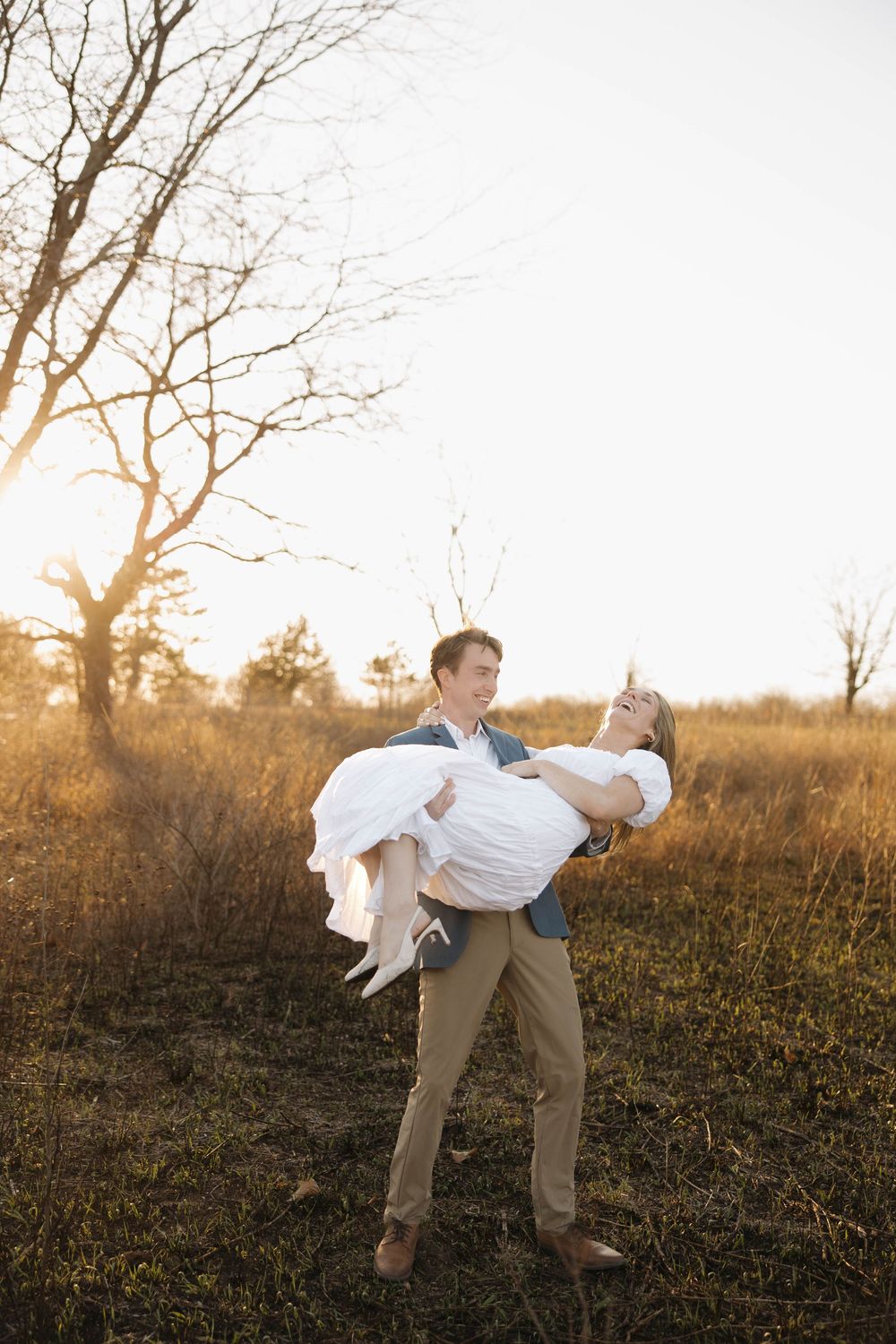 A couple shares a romantic moment at sunset in a field with bare trees and golden light filtering through the branches.