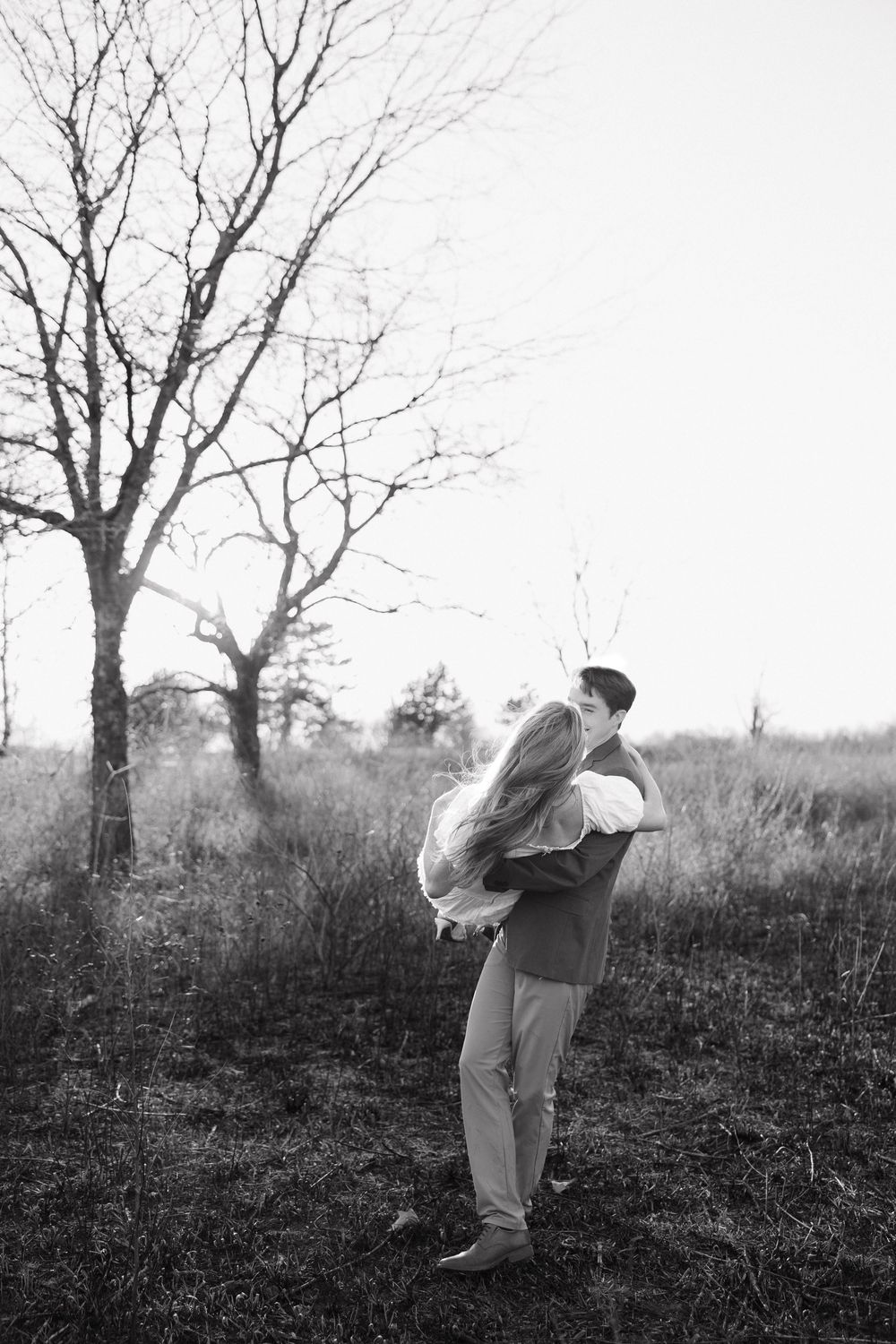 A black and white photograph shows a couple embracing in a field with bare winter trees in the background.