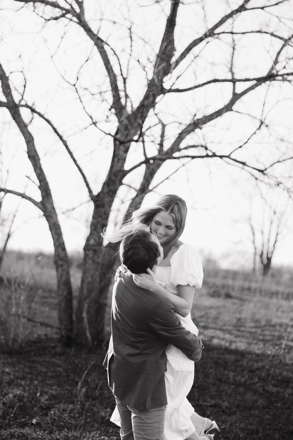 A romantic black and white photograph of a couple embracing outdoors under a bare winter tree.