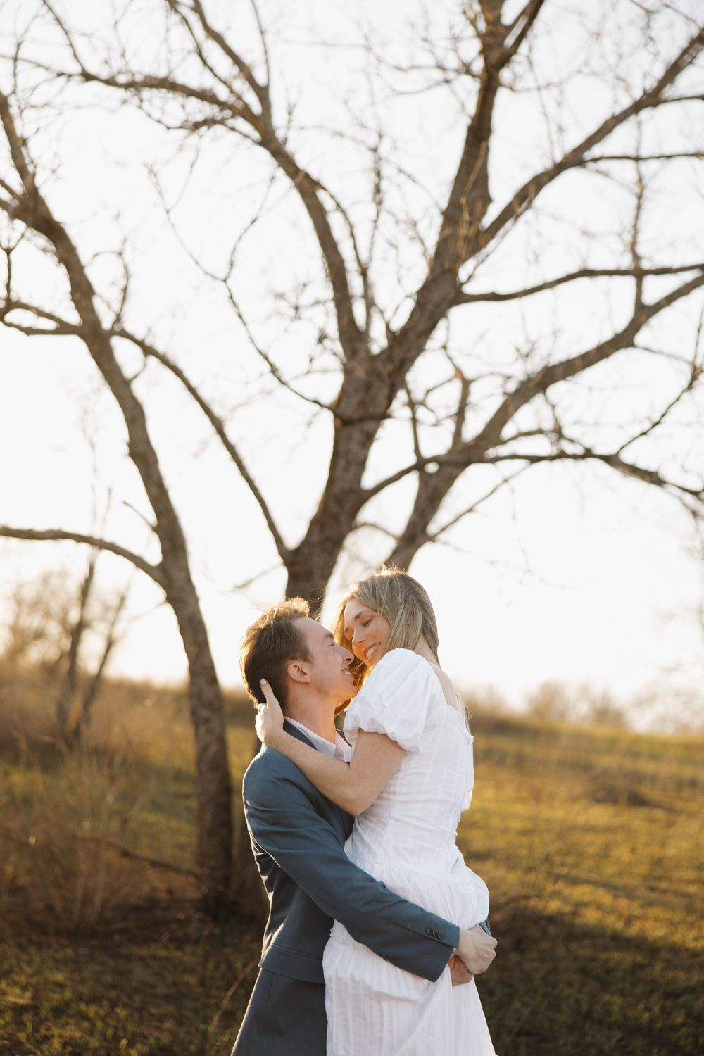 A romantic outdoor photo of a couple embracing in a natural setting with bare trees and golden sunlight.