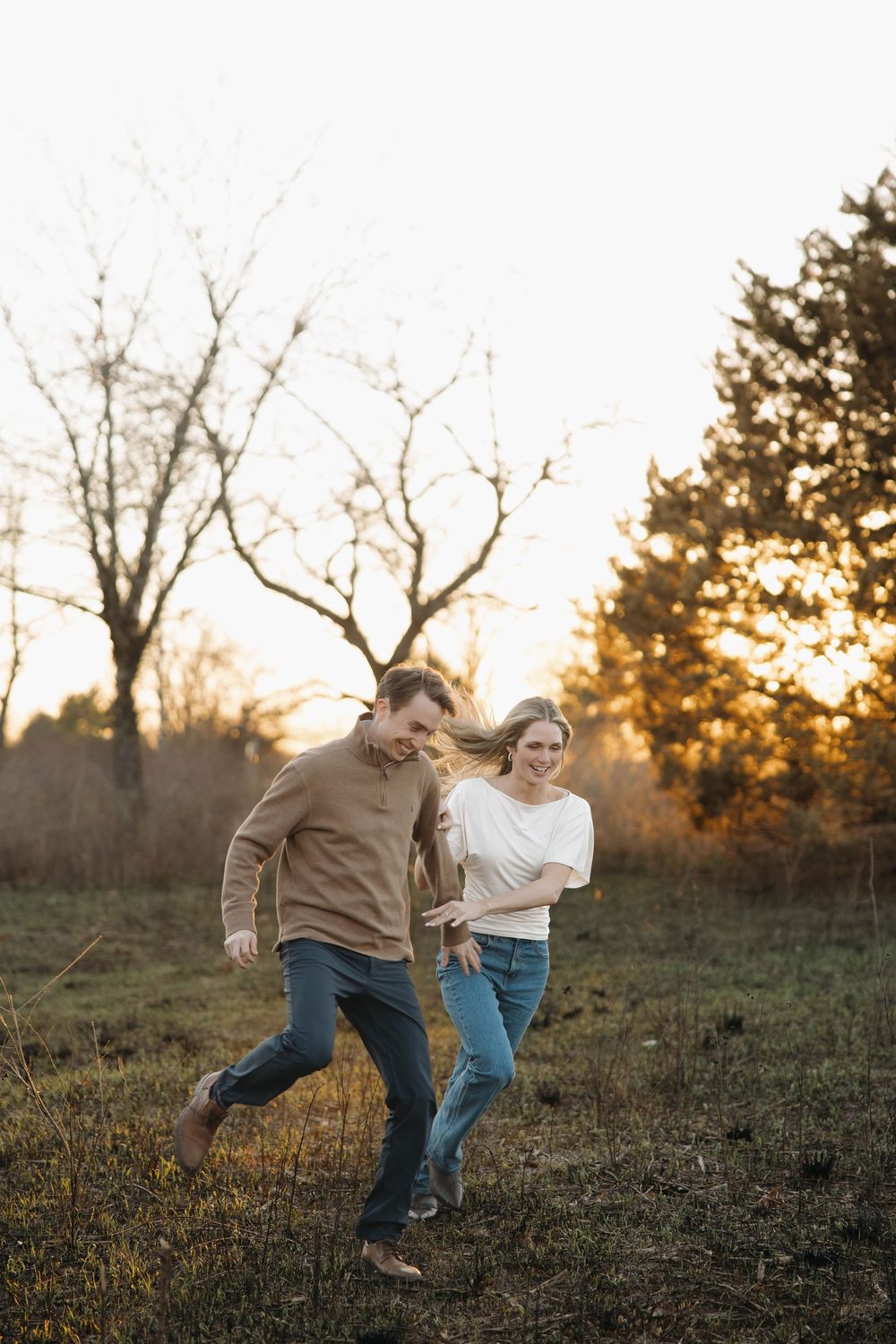 A couple runs playfully through a grassy field at sunset while holding hands and smiling.