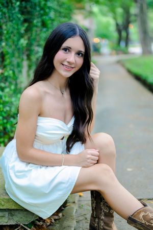 A figure in a white sundress poses outdoors in a lush green garden setting on a summer day.