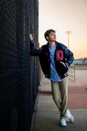 Student in varsity letterman jacket and plaid shirt poses against fence during sunset at athletic field.