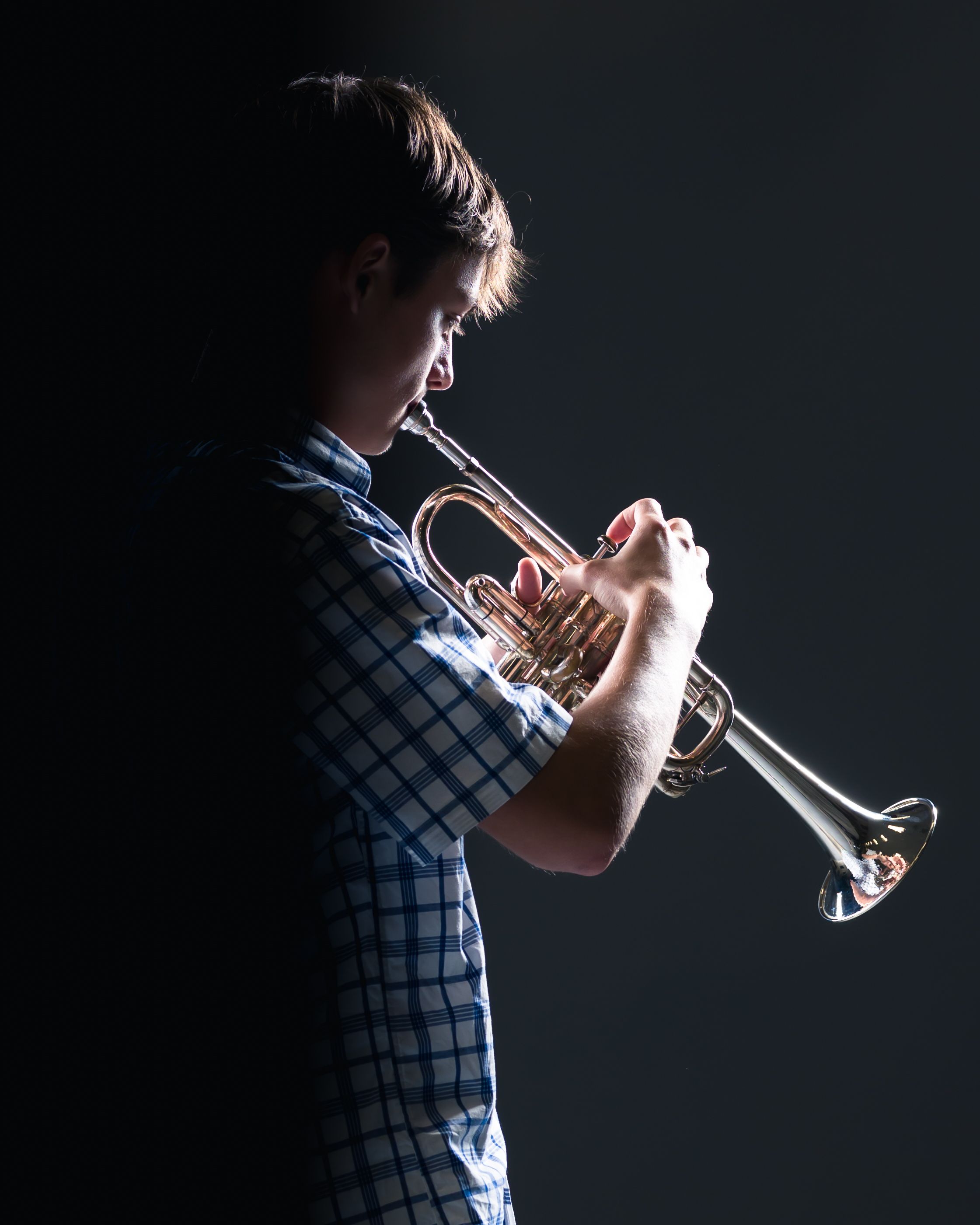 A musician plays trumpet in a checkered shirt against a dark background with dramatic side lighting.