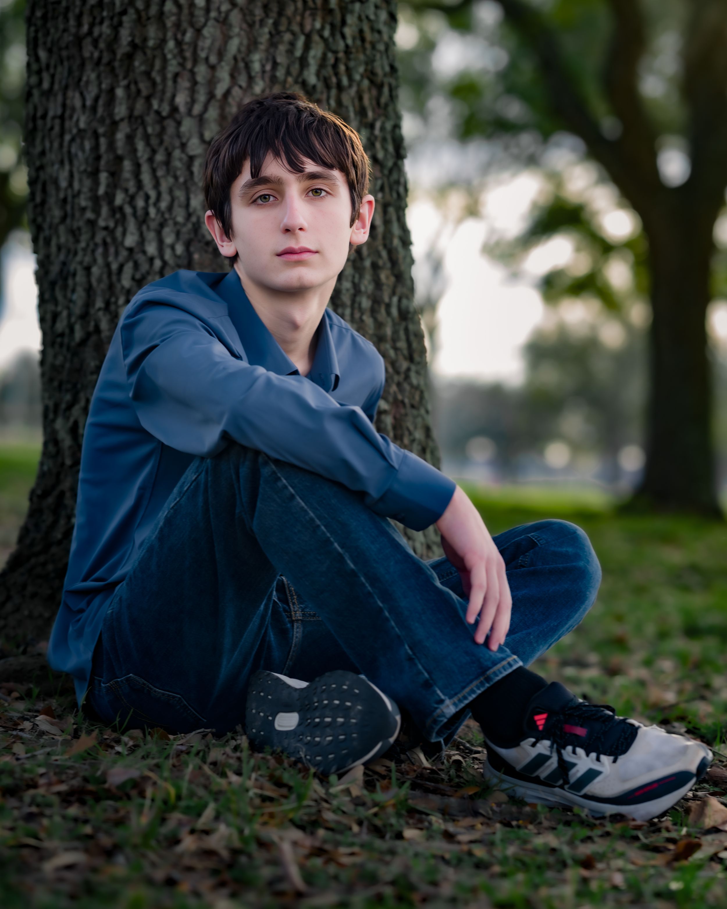A person in a blue hoodie sits against a tree trunk on fallen autumn leaves in a park setting.