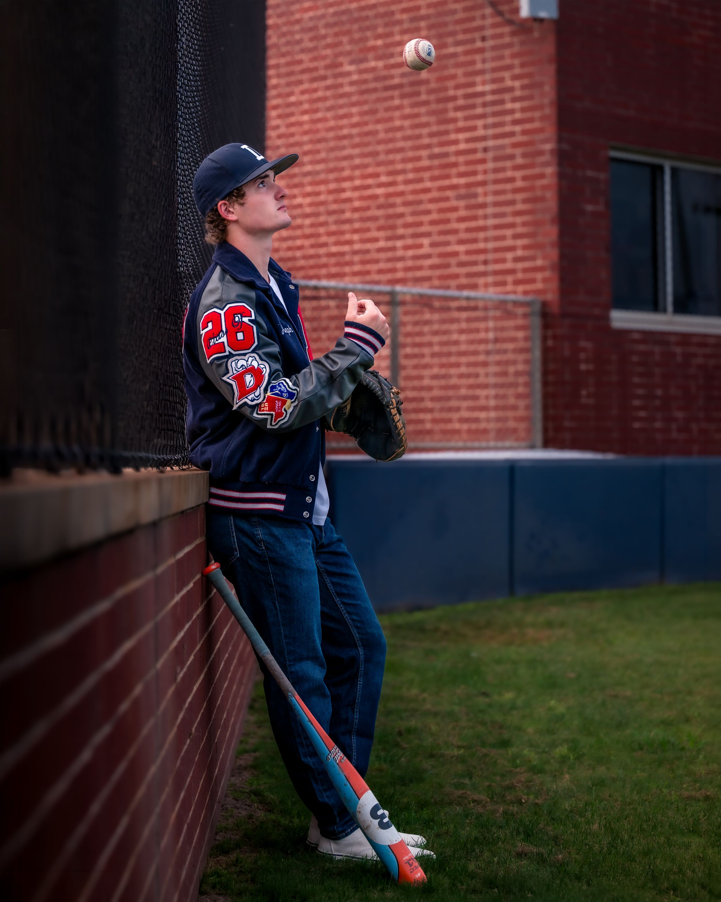 Young baseball player in navy jacket number 26 tossing a ball while leaning against a brick wall with glove in hand.