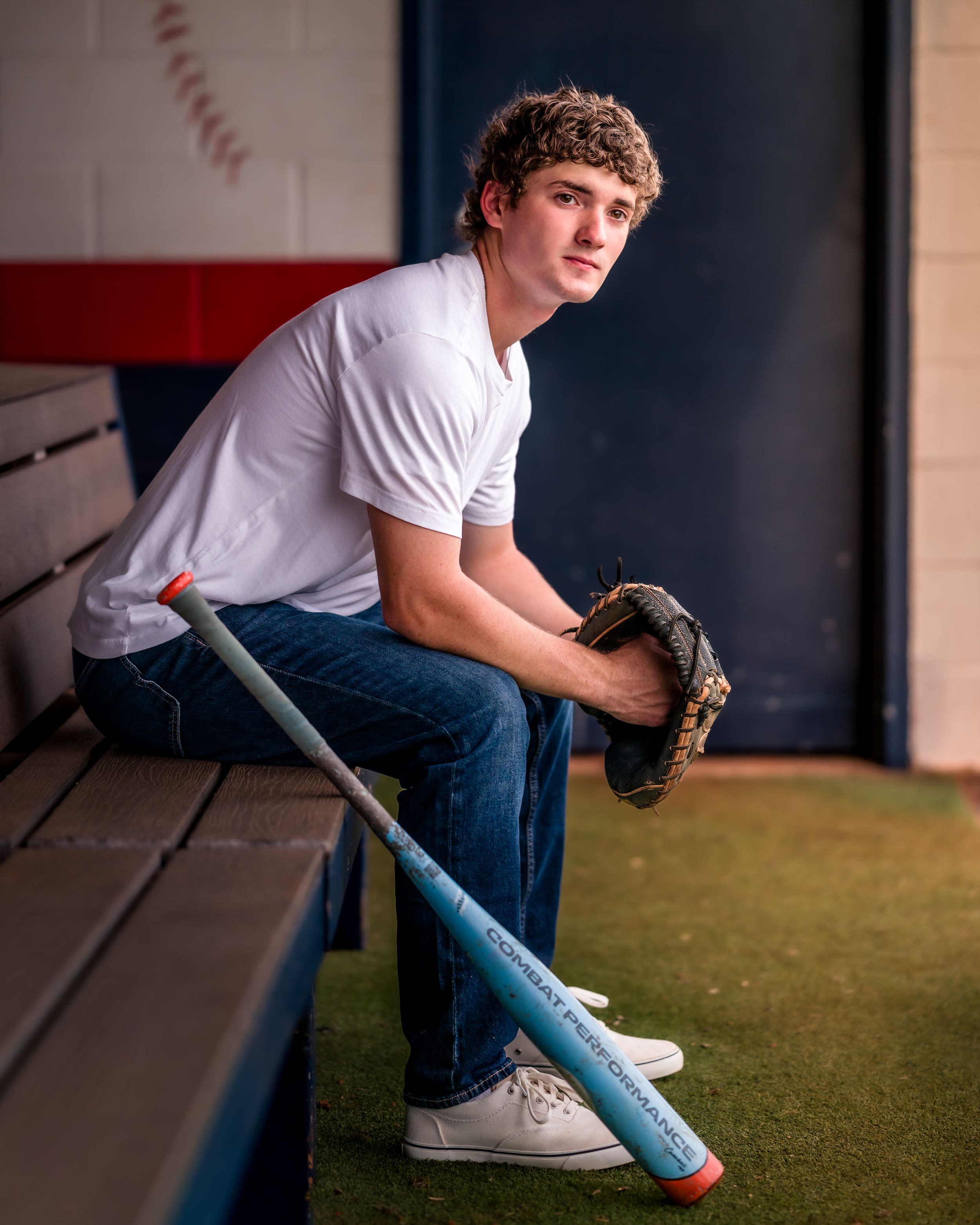 Teen baseball player sits in dugout holding bat and glove, wearing white tee and jeans, in a professional senior portrait.