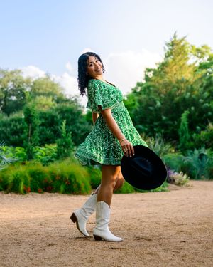A person wearing a green floral dress and black pants performs dynamic dance moves on a dirt path at sunset.