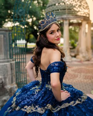 A person in a stunning royal blue and gold quinceañera dress poses elegantly in an outdoor garden setting at sunset.