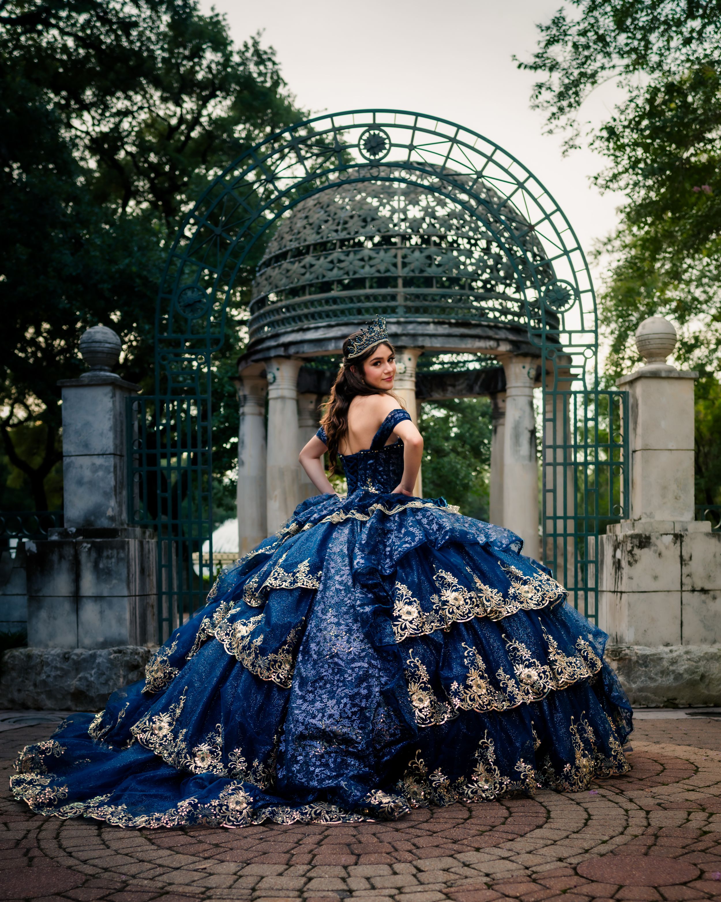 A stunning quinceañera photoshoot sequence in a navy blue and gold ballgown against an ornate garden entrance gate.