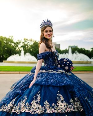A quinceañera dress in navy blue with intricate gold embroidery details photographed in an outdoor setting at sunset.