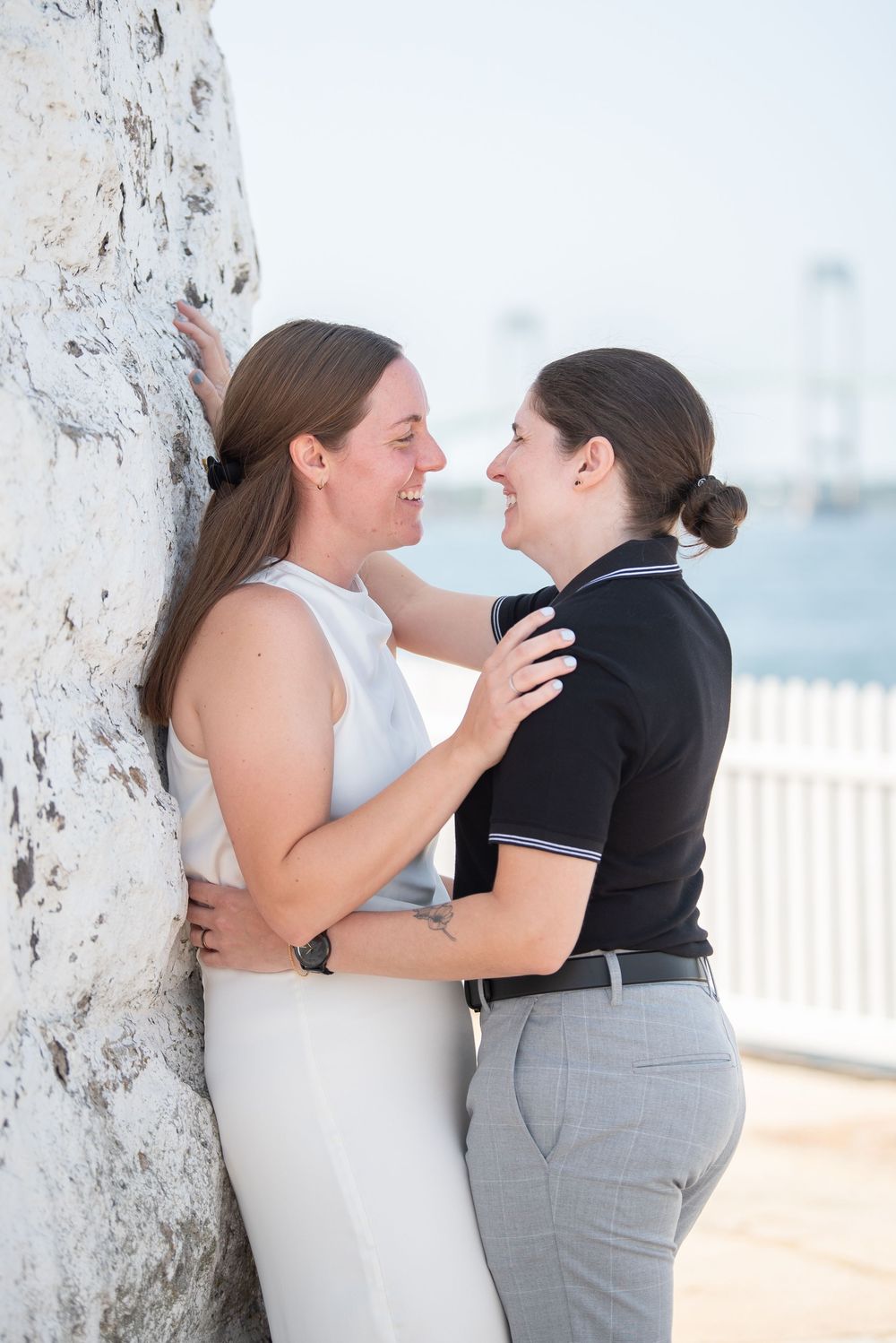 Two people in contrasting outfits embrace against a white pillar with ocean views.
