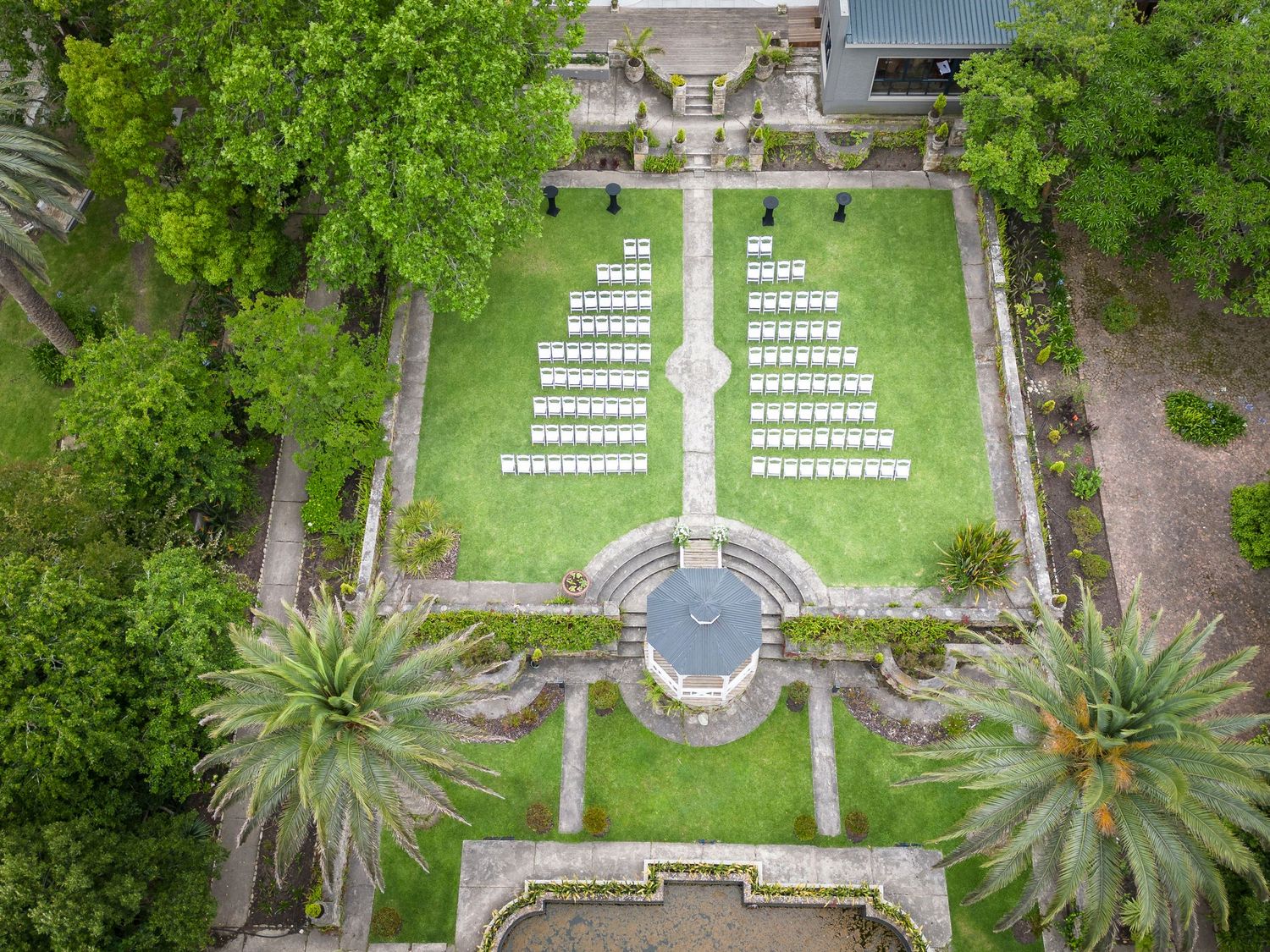 Aerial view of a formal garden venue with white chairs arranged for an outdoor wedding ceremony, surrounded by lush greenery.