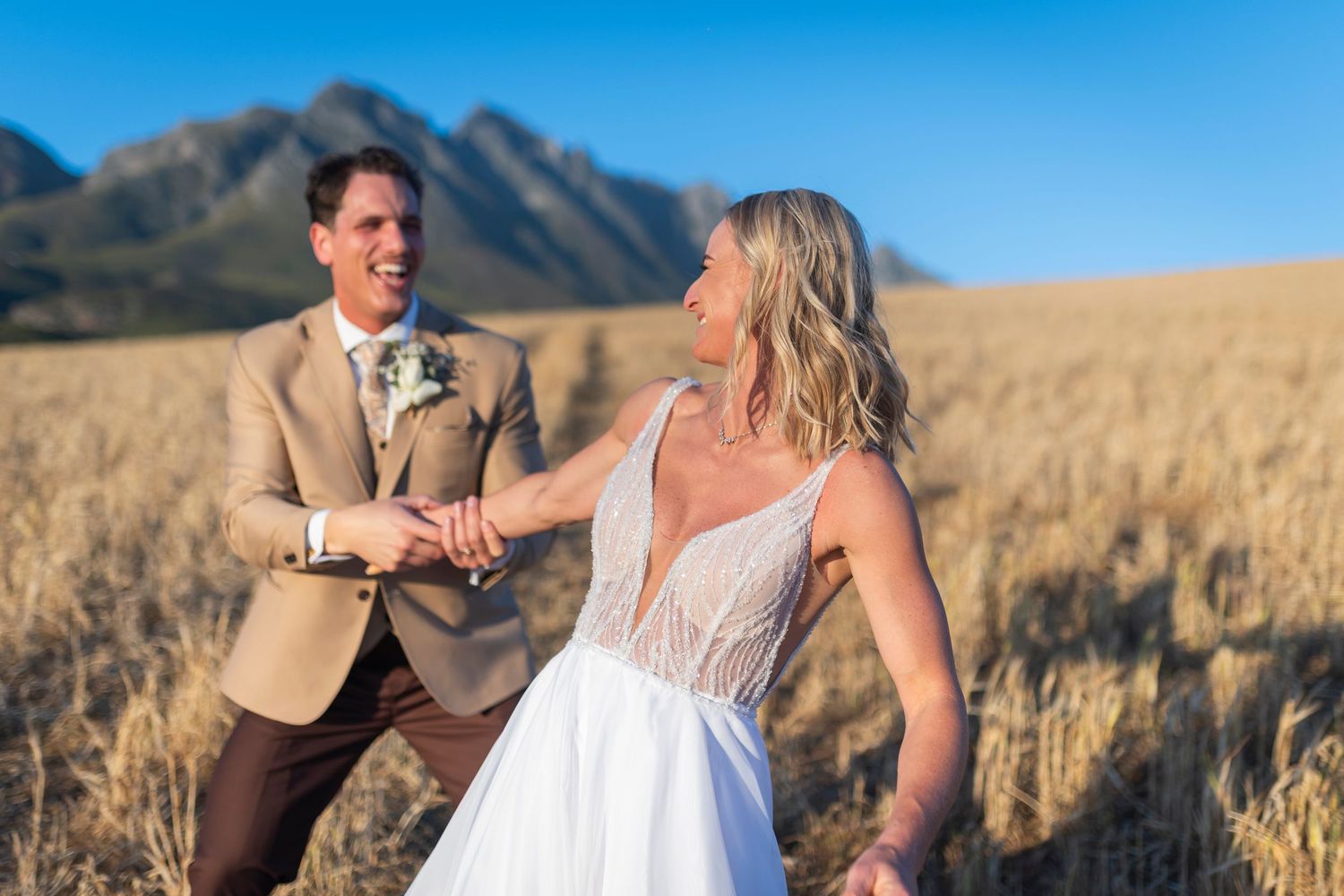Bride and groom holding hands in golden wheat field with mountain backdrop on their wedding day.