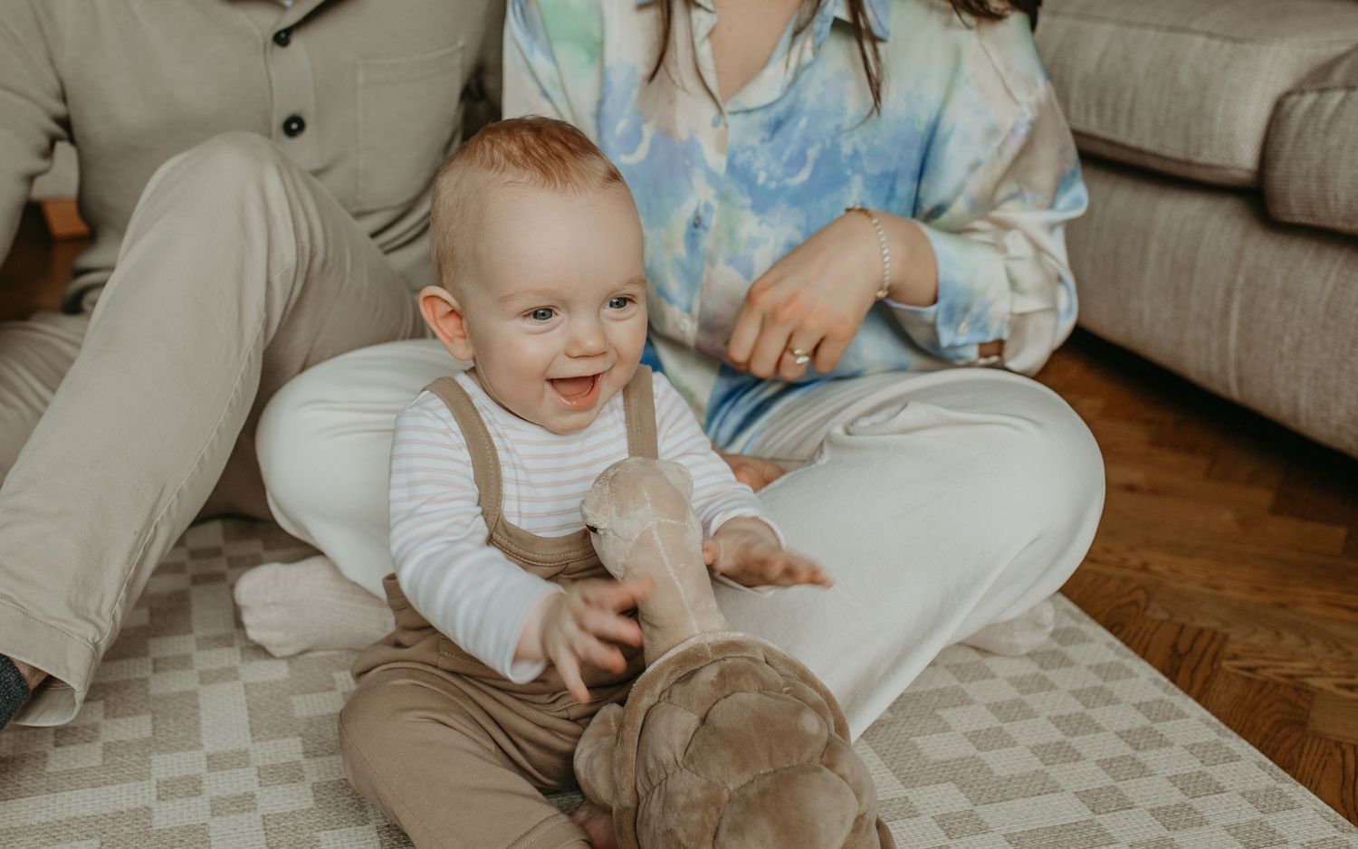A smiling baby in white overalls plays on a carpeted floor while sitting with someone on a couch.