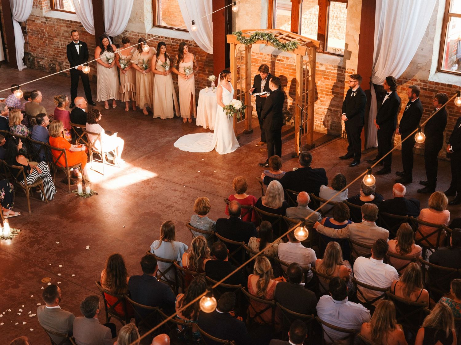Wedding ceremony in a rustic brick venue with white drapes and guests seated in rows watching the ceremony.
