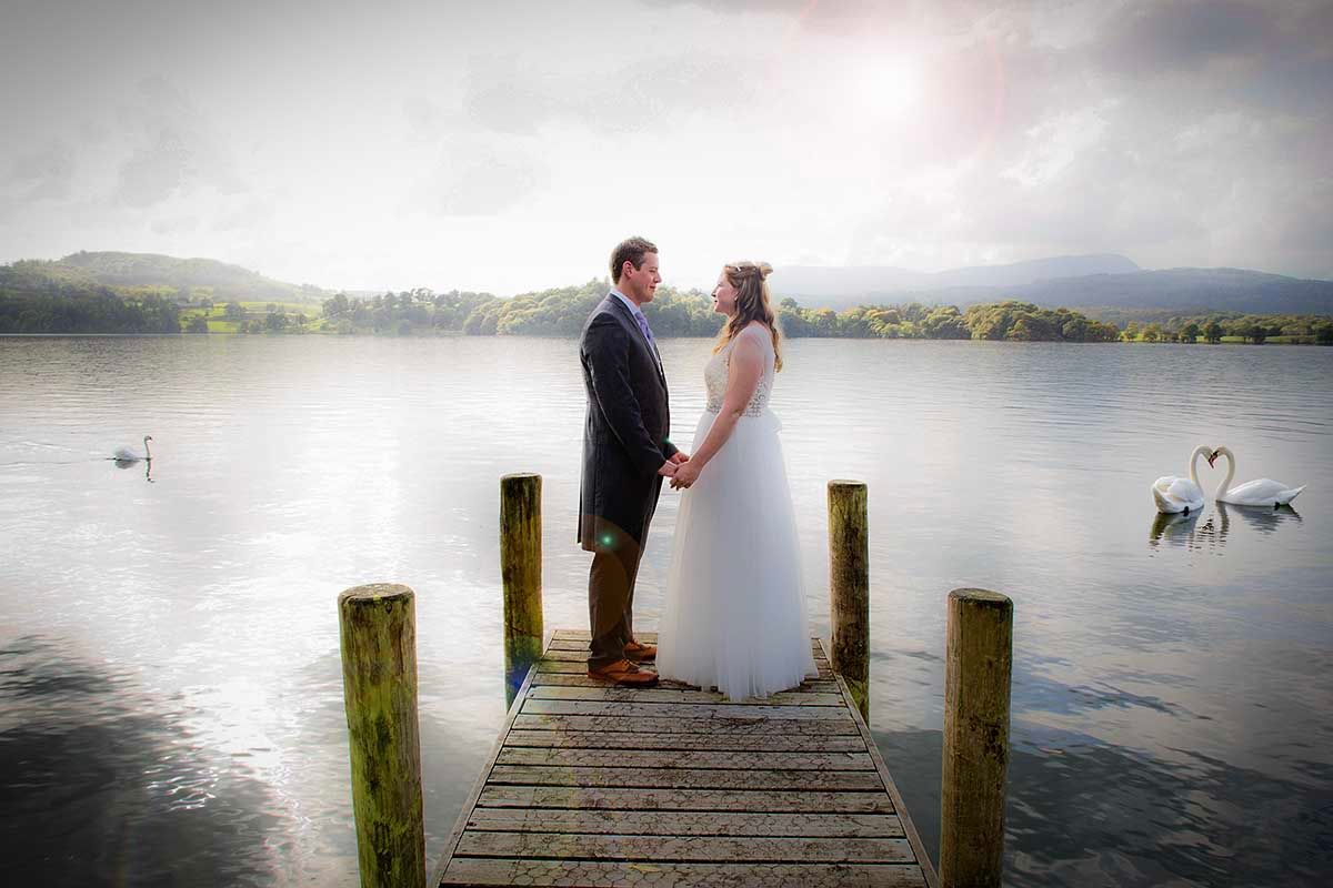 Bride and groom holding hands on a wooden dock by a serene lake with swans, sharing a romantic moment on their wedding day.