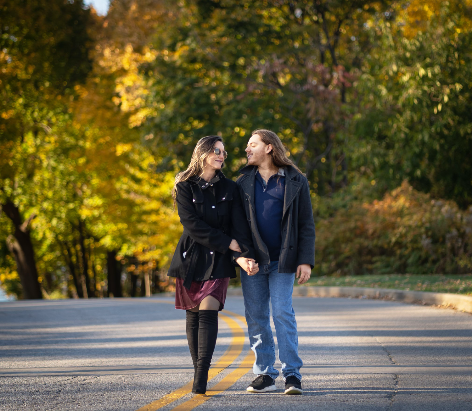 Two people walk together on a paved path surrounded by autumn trees with golden foliage in a park setting.