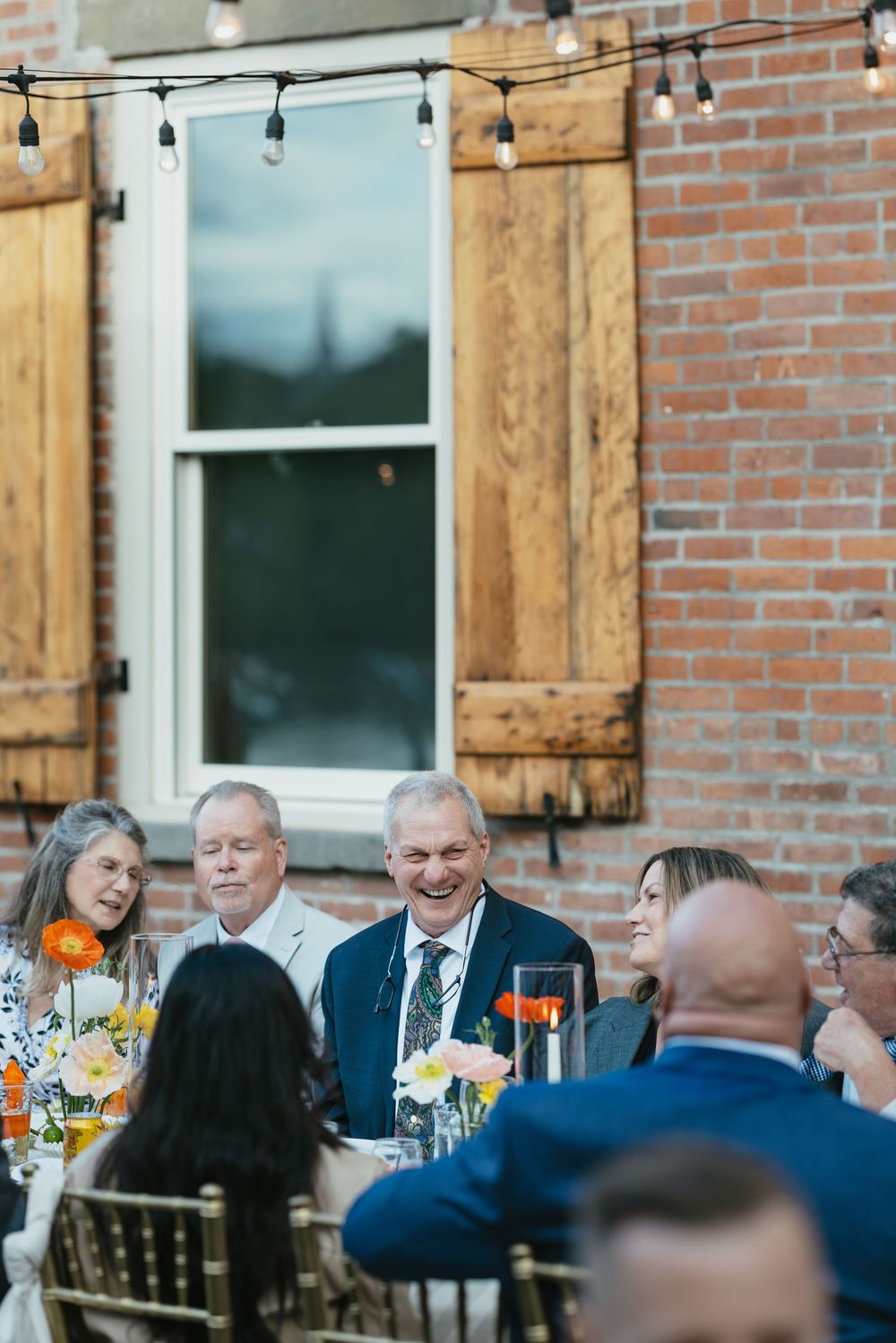 Guests seated at an outdoor dinner party under string lights next to a brick building with wooden shutters.