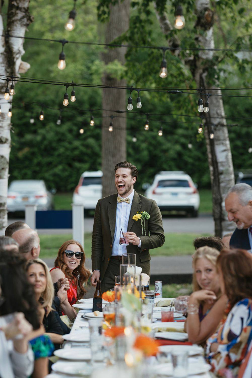Wedding guests enjoy an outdoor reception dinner under string lights and birch trees at dusk.