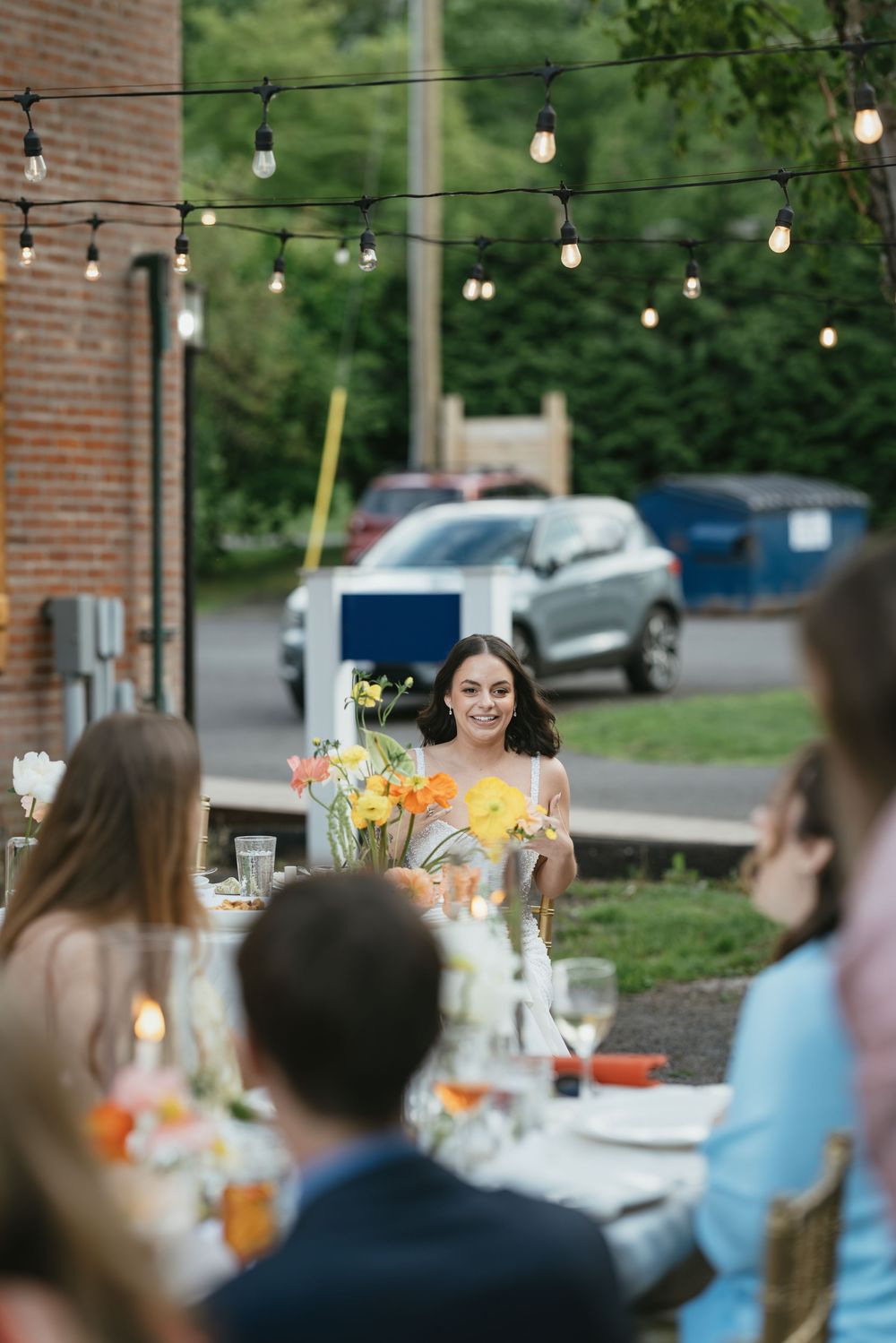 People enjoying conversation at a long outdoor dining table with string lights and floral centerpieces.