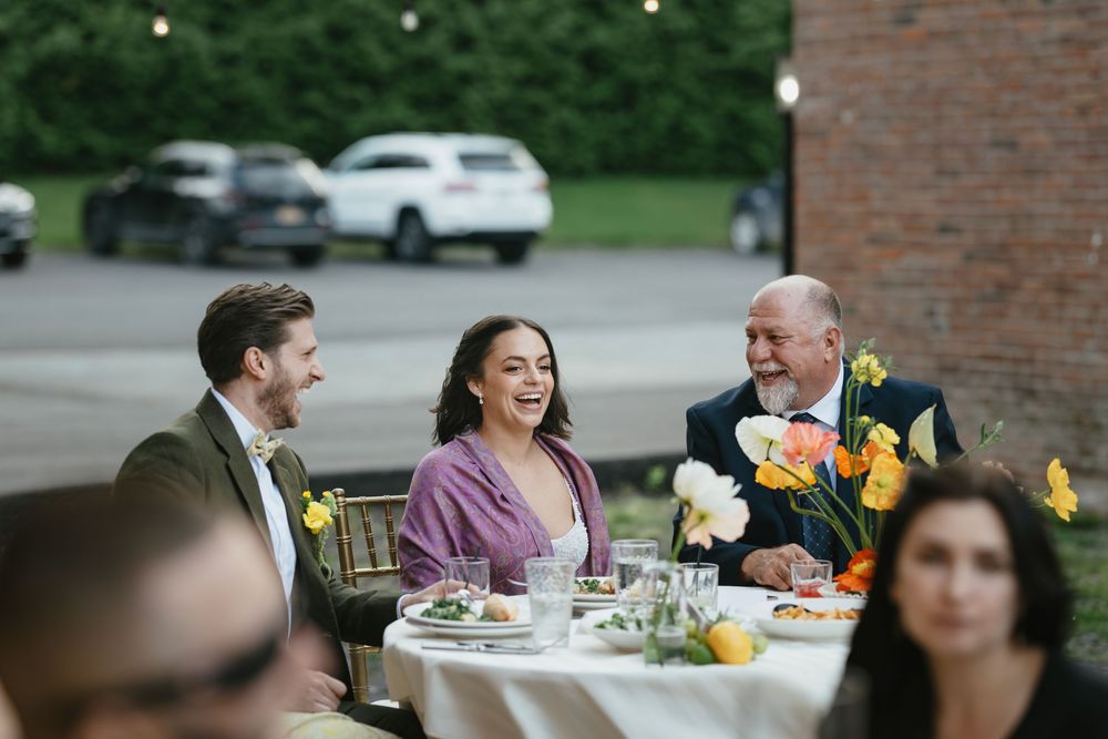 Wedding reception guests seated at tables with floral centerpieces against a brick wall backdrop.