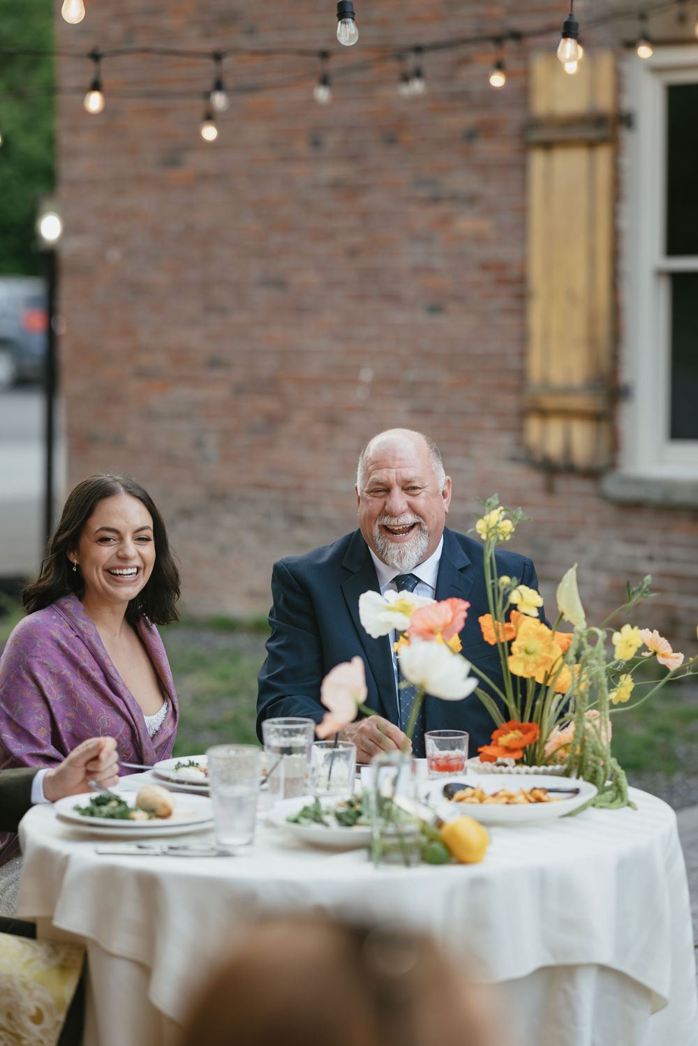 Guests enjoying dinner at round tables decorated with colorful floral arrangements under string lights.