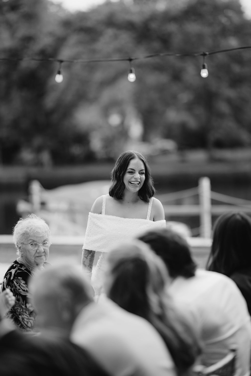 Black and white photo of wedding guests enjoying an outdoor reception under string lights.