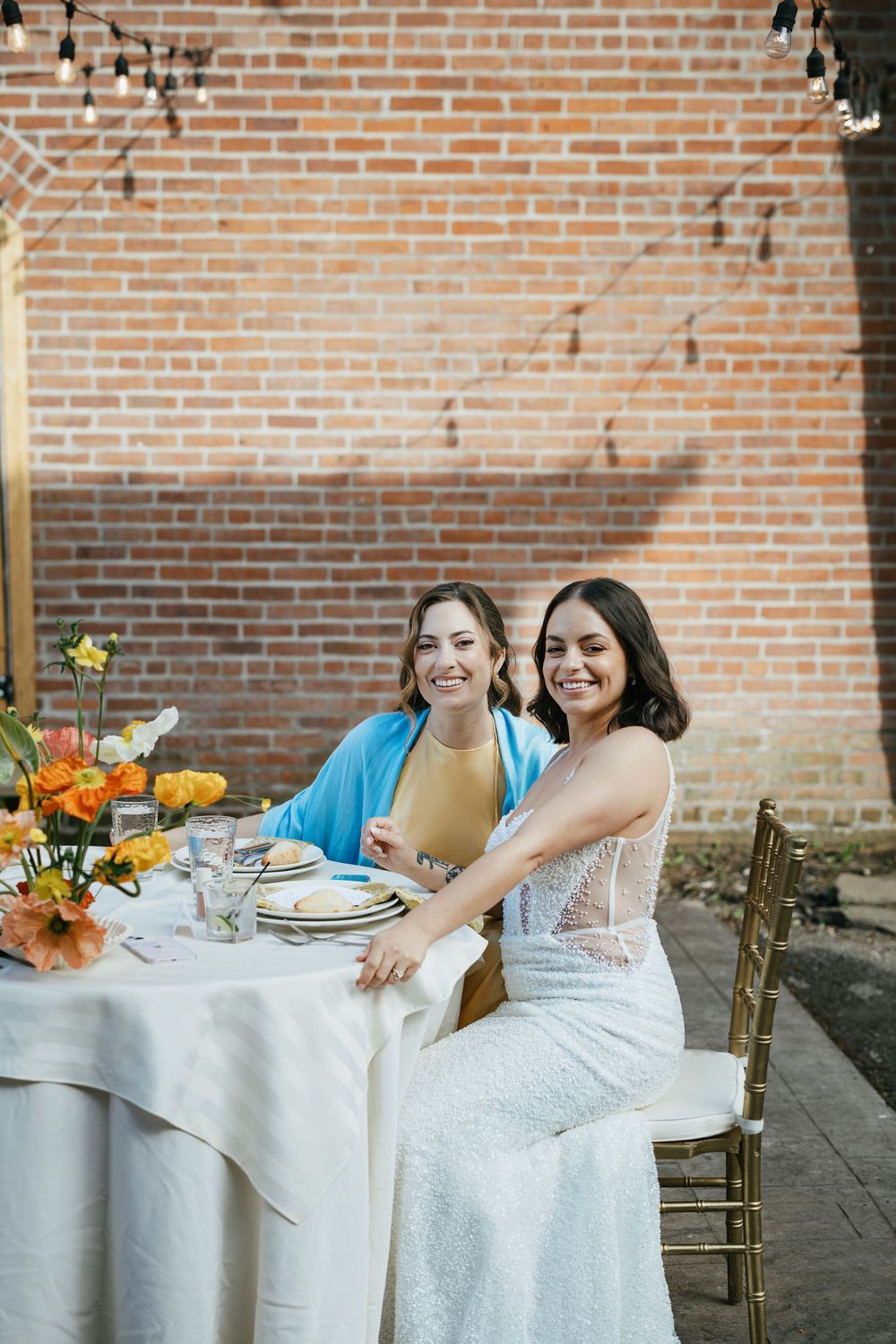 Three white-gowned figures seated at an elegant outdoor table setting with orange floral arrangements against a brick wall.