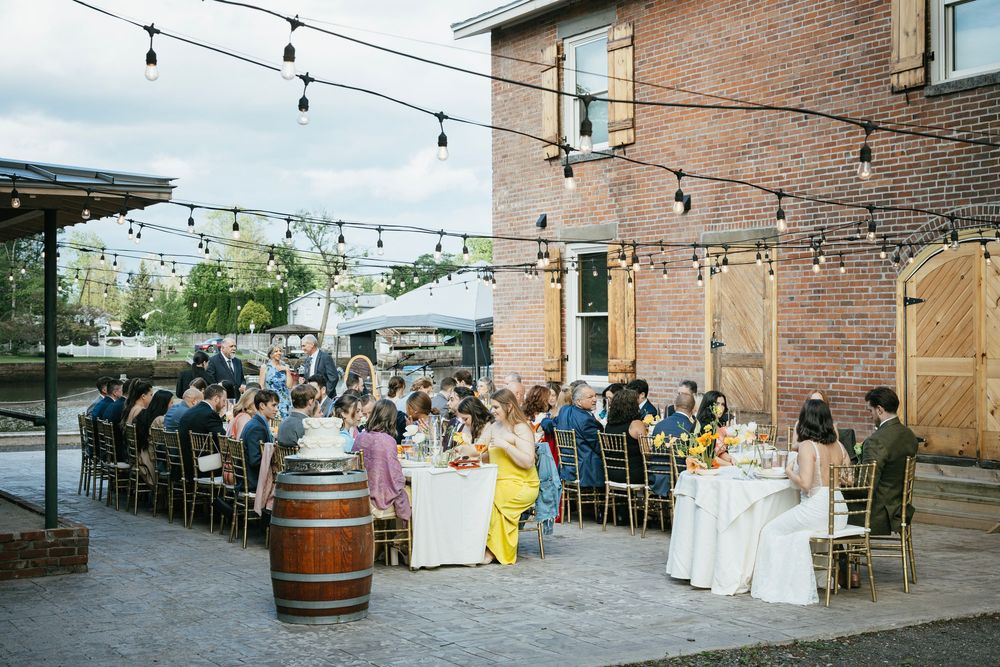 String lights illuminate an outdoor brick patio dining area with wine barrels and tables set for guests.