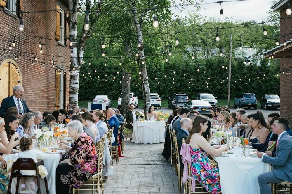 Wedding guests gather at a rustic outdoor reception venue with string lights and brick walls creating an intimate atmosphere.