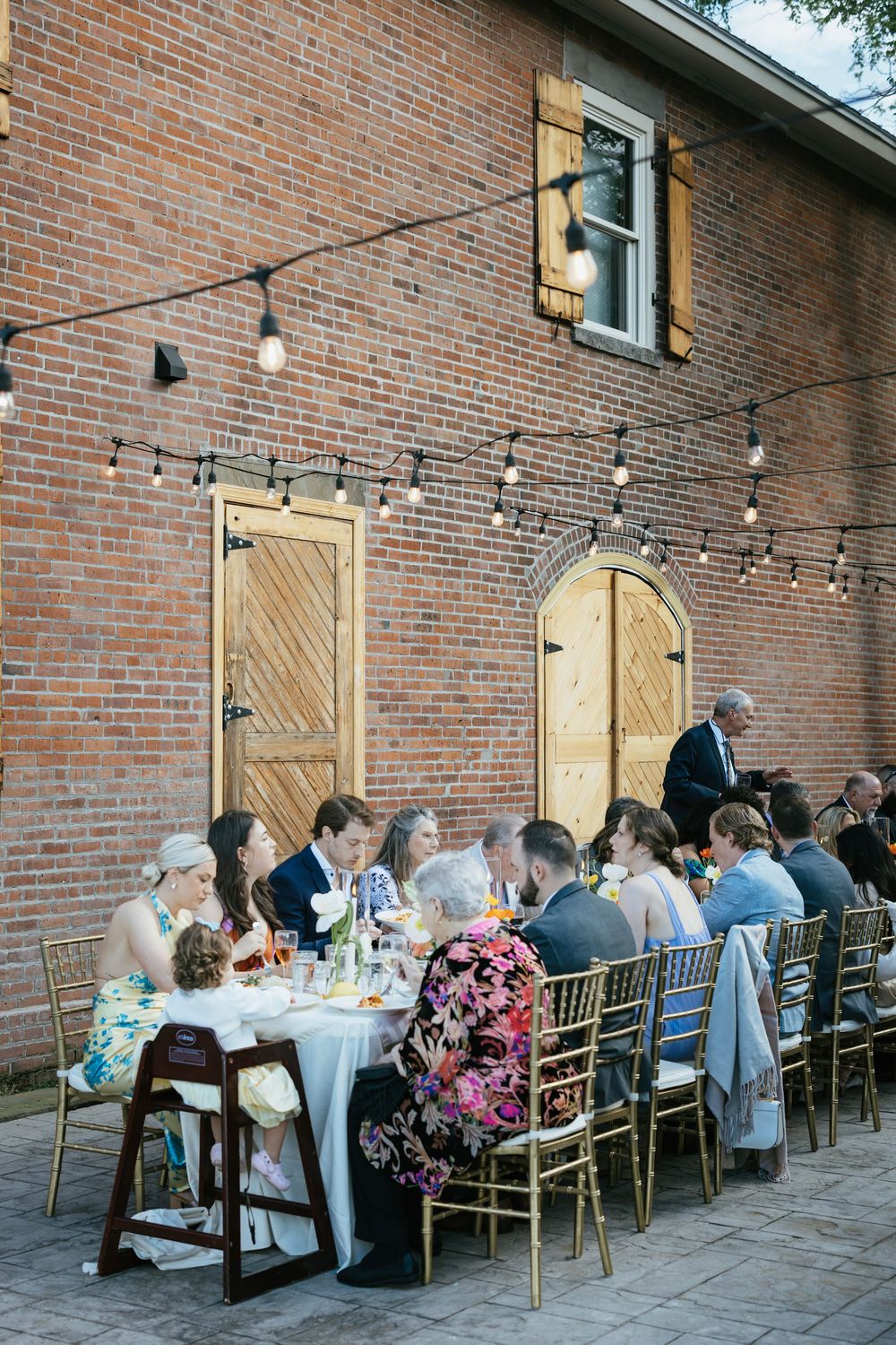 Guests seated at elegant outdoor dining tables along a brick wall with string lights creating an intimate atmosphere.