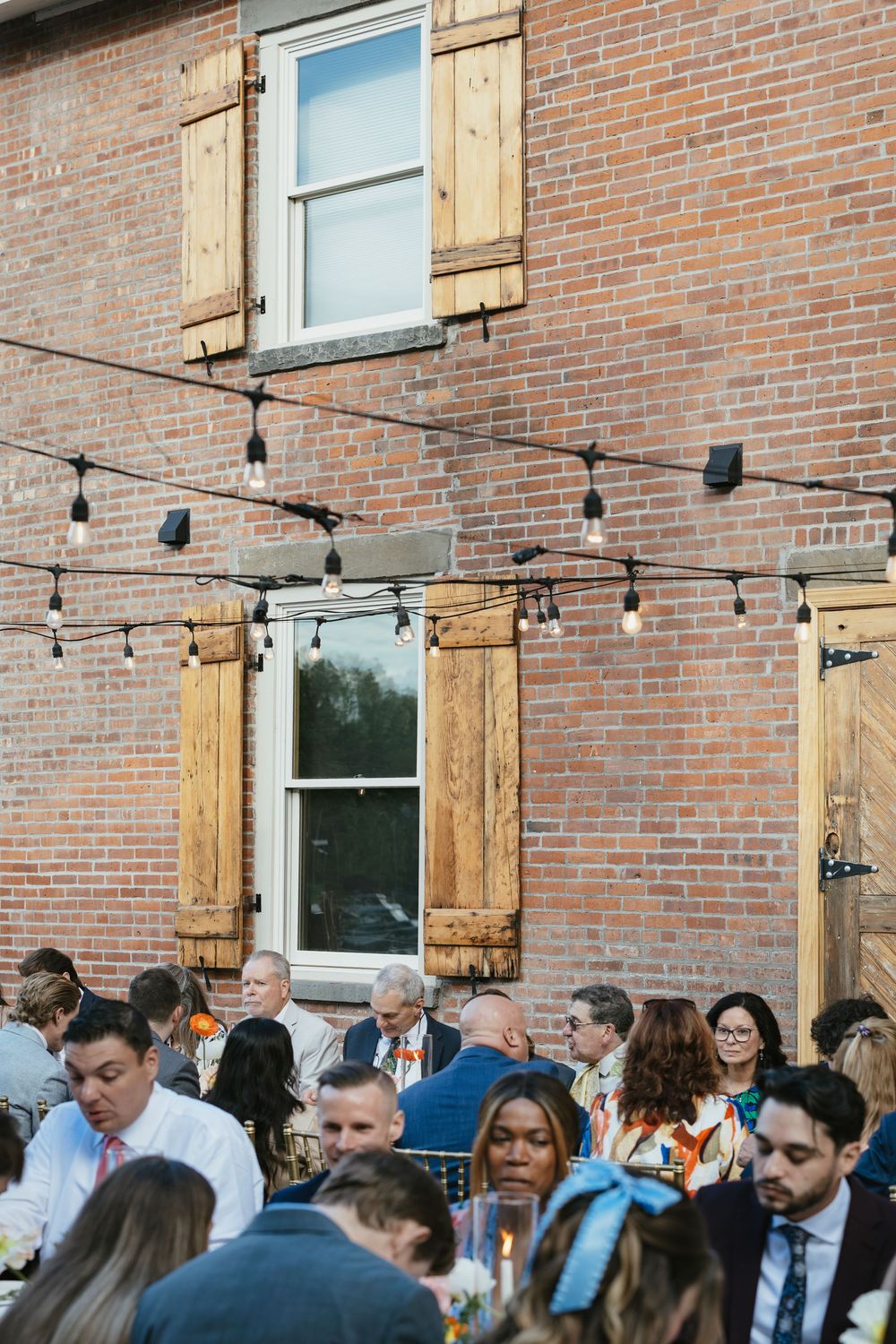 Wedding guests mingle outdoors at a rustic brick venue with string lights overhead during a reception celebration.