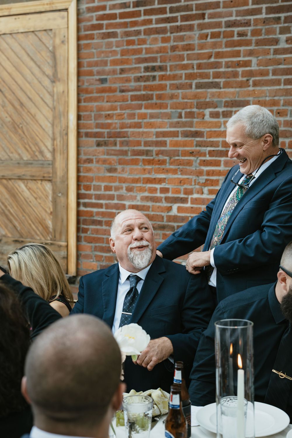 Guests enjoying conversation and laughter at a formal dinner event with brick wall backdrop.