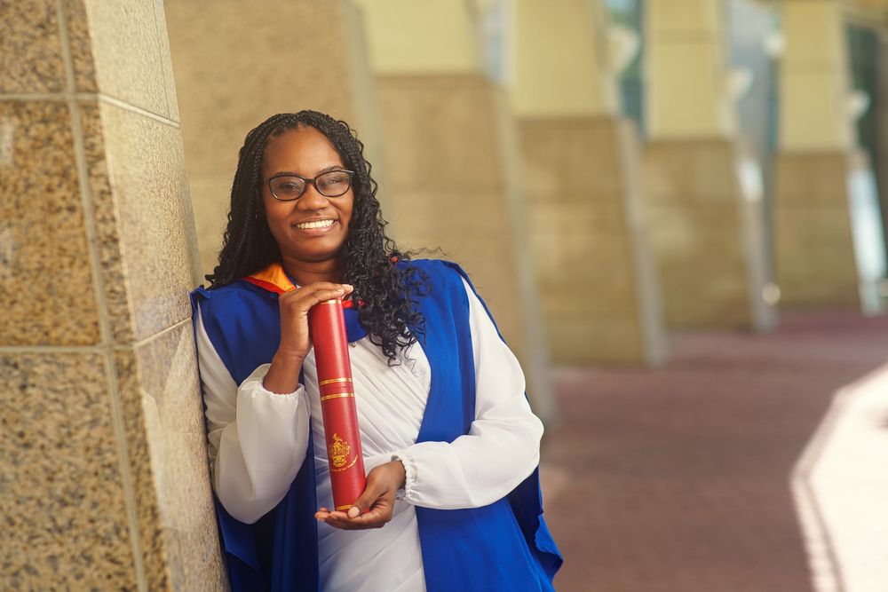A student in graduation regalia holds a red diploma case while leaning against a stone column outdoors.