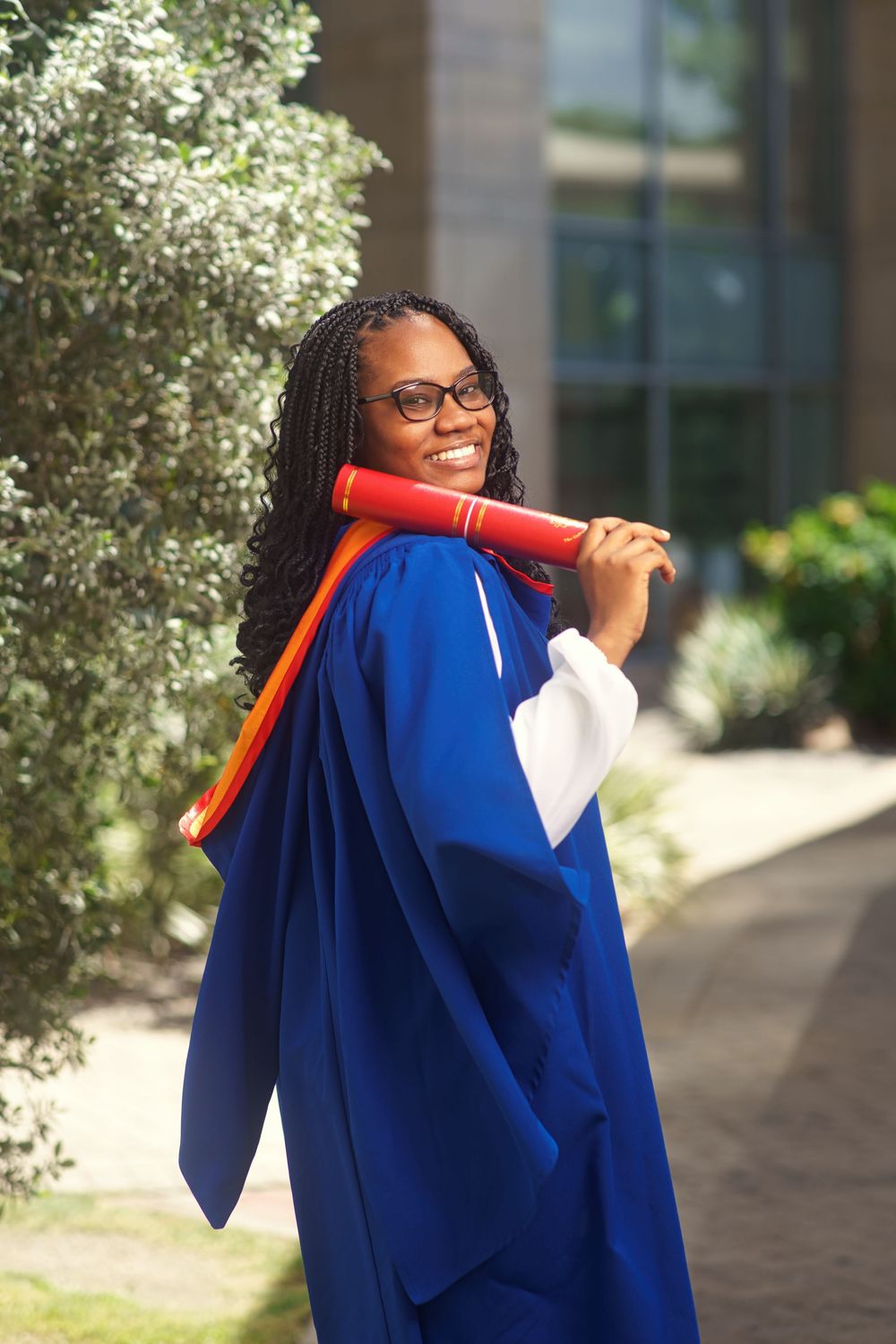 Graduate in blue academic regalia poses with red diploma holder on sunny campus walkway.