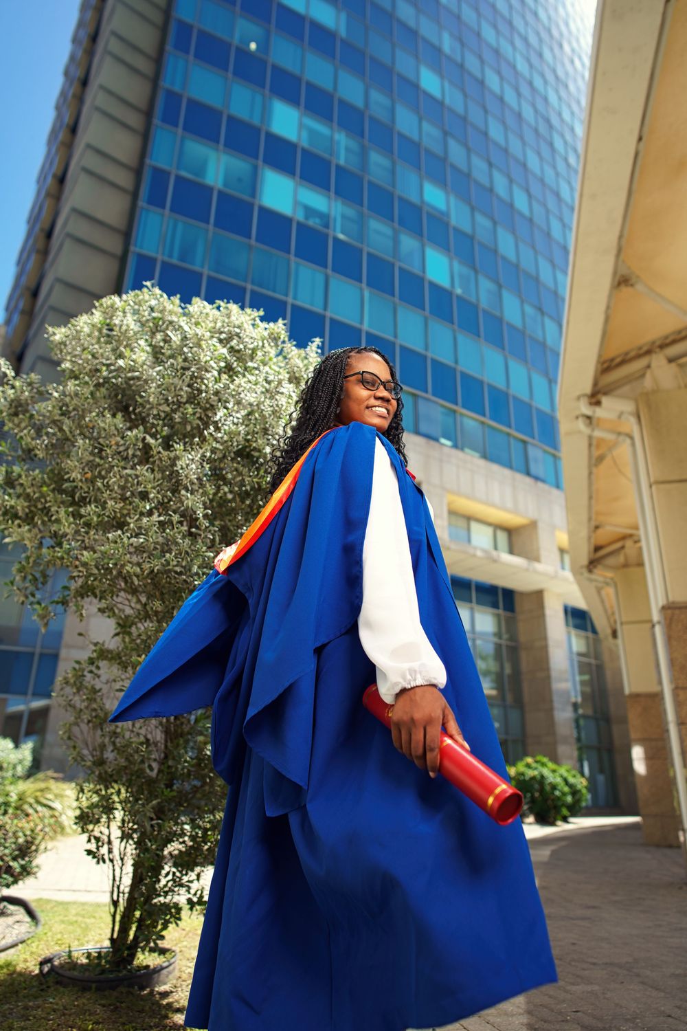 Graduate in royal blue graduation gown stands in front of modern glass building with olive tree in background.