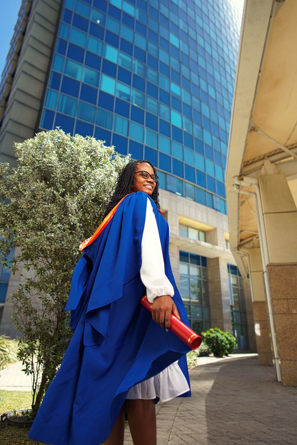 Graduate in blue academic regalia poses against modern glass building exterior with columns and olive trees.