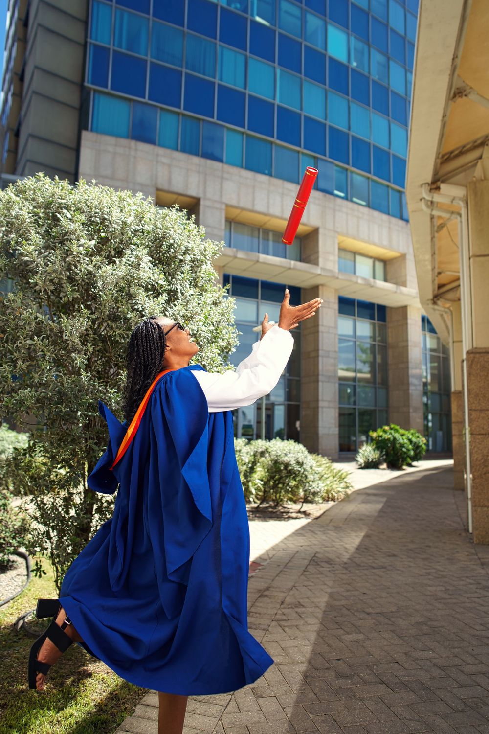 Graduate in blue academic regalia poses triumphantly against modern glass building on graduation day.