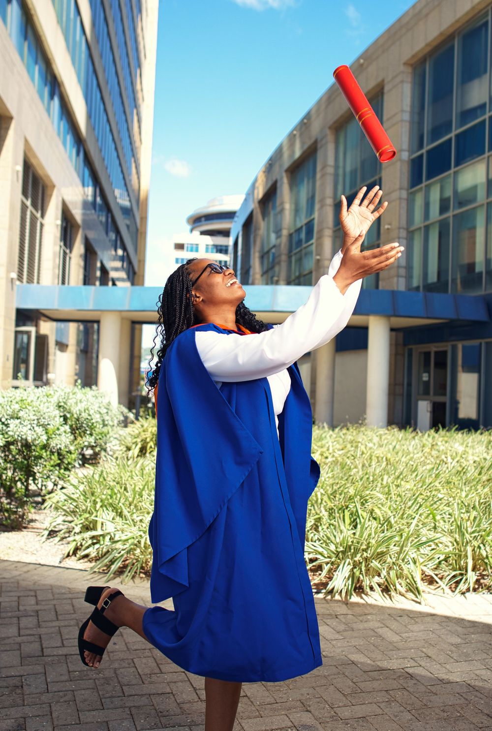 A person in a bright blue graduation gown performs a series of celebratory dance moves outside a modern building.