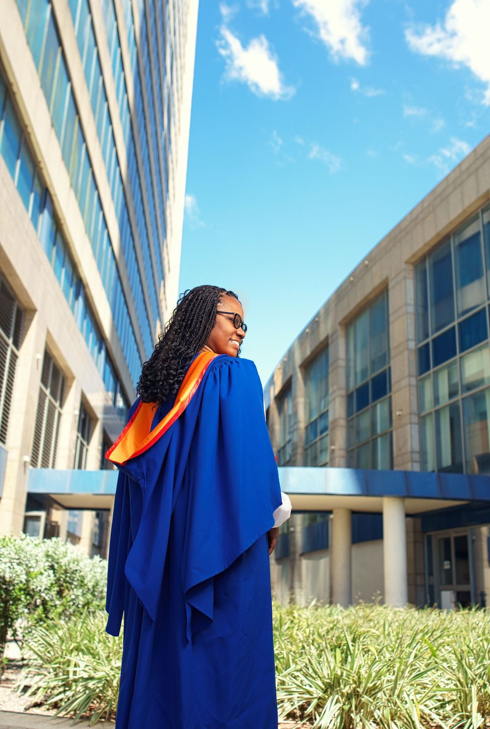 A graduate in royal blue regalia strikes joyful poses during a photoshoot on a sunny day near campus buildings.