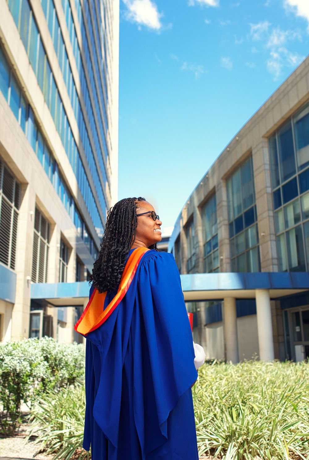 Graduate celebrates achievement in blue cap and gown outside modern university building under bright blue sky.