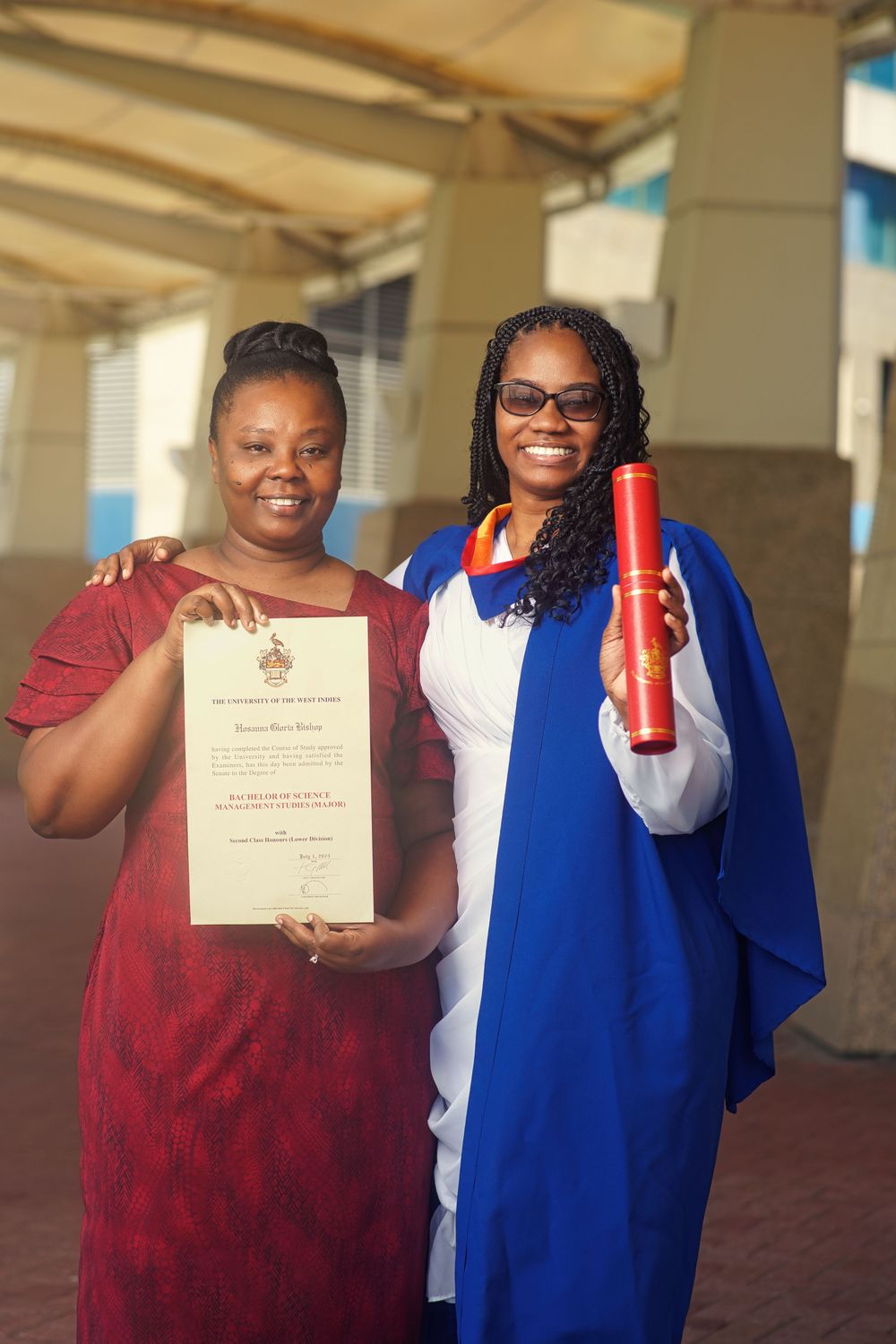 Two graduates celebrate with diploma and red holder while wearing academic regalia outdoors.